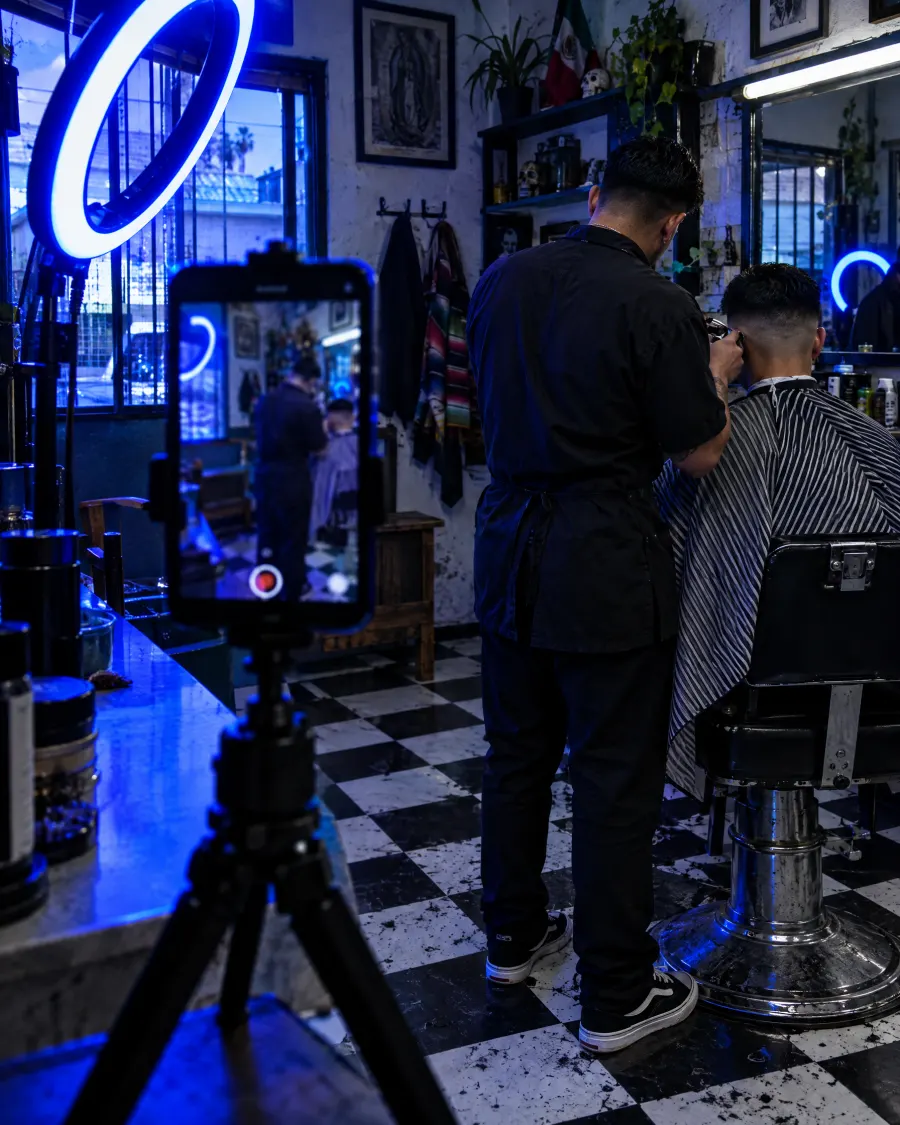 Barbero cortando el pelo mientras un celular en un trípode graba la escena con luz azul.